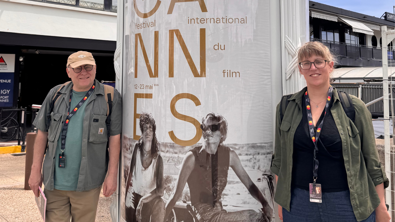 Two people, from left to right: Mark Cosgrove and Maddy Probst stand in front of a large Cannes Film Festival poster outdoors, both wearing badges and casual clothing.