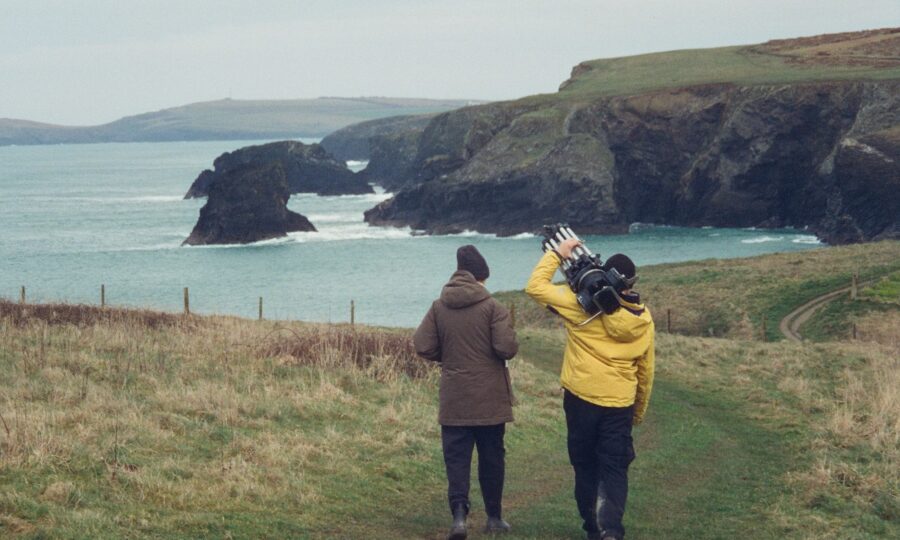 Two people walk along a grassy cliff path, holding a tripod and camera, with a rocky coastline with ocean waves in the distance.