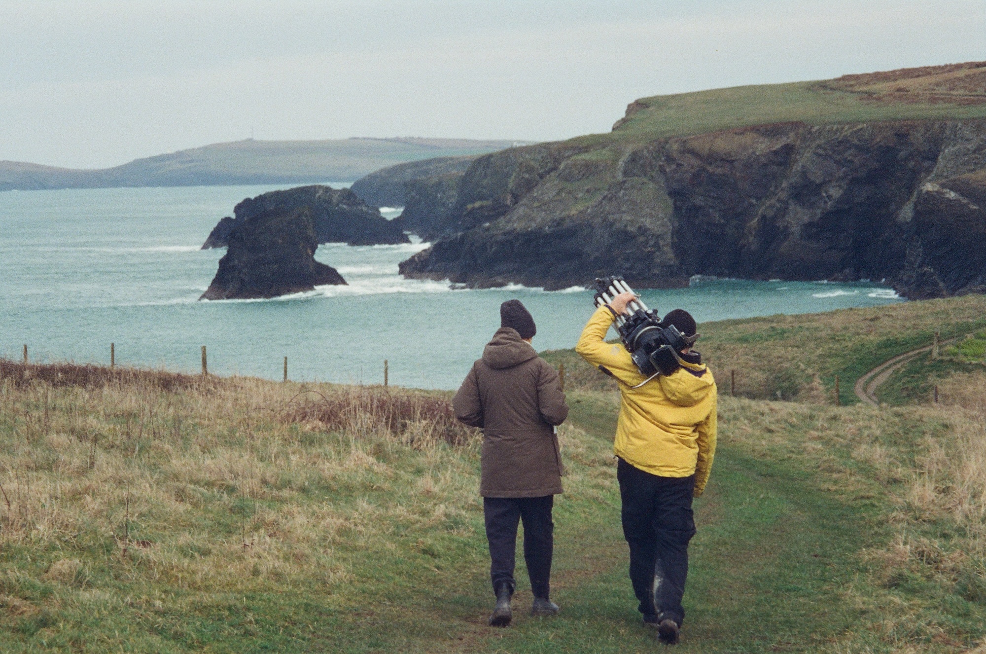 Two people walk along a grassy cliff path, holding a tripod and camera, with a rocky coastline with ocean waves in the distance.