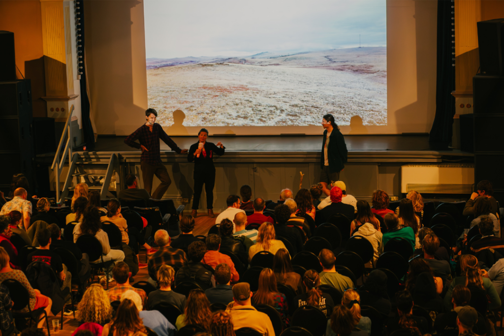 Three presenters stand in-front of a large projector speaking to a full cinema audience.