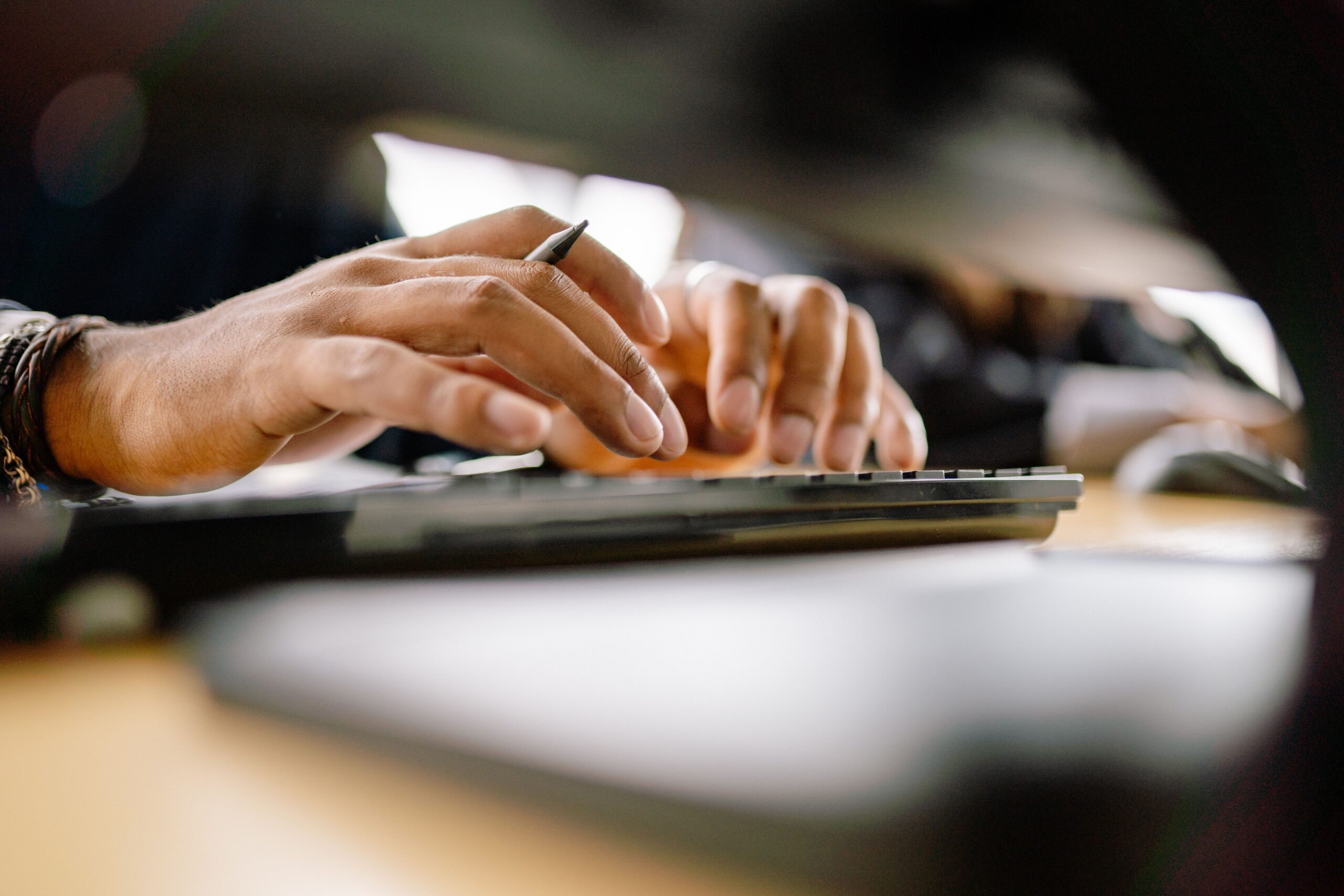 Close-up of a person’s hands typing on a computer keyboard, with a blurred background and a pen held between fingers.