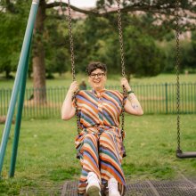 A woman with short hair and glasses wearing a multi coloured, stripey outfit sits on a swing smiling,  with trees and grass in background.