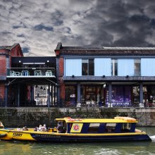 Exterior shot of Watershed building with a cloudy sky and a blue and yellow Bristol Ferry Boat passing on the water.  