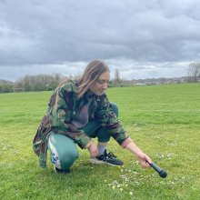 Person crouching down on the grass holding a microphone to the ground. 