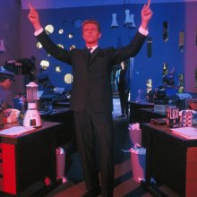 Daivd Bowie pointing his hands in the air standing amongst desks.