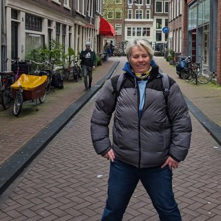 A full body photo of a white woman in jeans and a puffer jacket, hands on hips in an Amsterdam street, looking into the camera