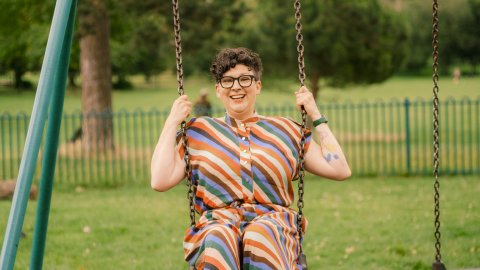 A woman with short hair and glasses wearing a multi coloured, stripey outfit sits on a swing smiling,  with trees and grass in background.