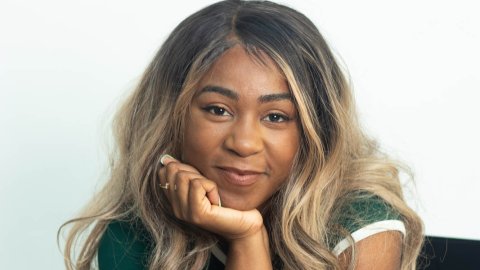 A young Black woman sits with a hand under her chin, she has long blonde hair and smiles softly into the camera.
