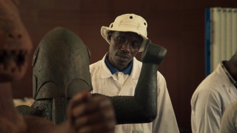 A man wearing a white bucket hat cocks his head as he looks at a masked statue.