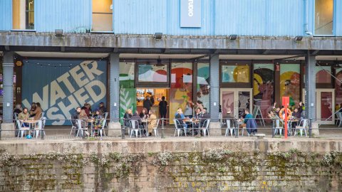 Watershed's blue exterior. Six tables of people sit outside Undershed bar enjoying drinks.