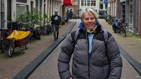 A full body photo of a white woman in jeans and a puffer jacket, hands on hips in an Amsterdam street, looking into the camera