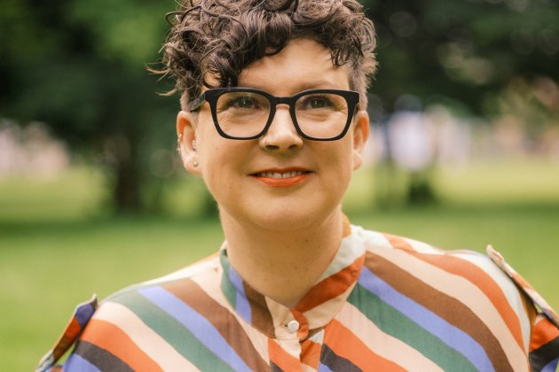 Head and shoulders of a woman with short hair and glasses wearing a multi coloured, stripey top with trees in background.
